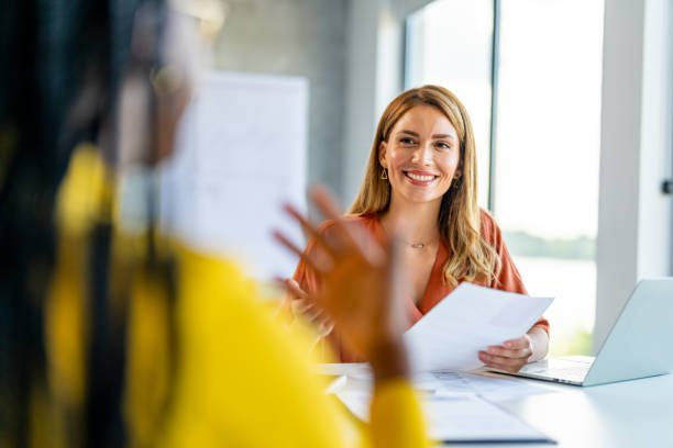 Smiling woman in an office setting holding papers during a meeting.